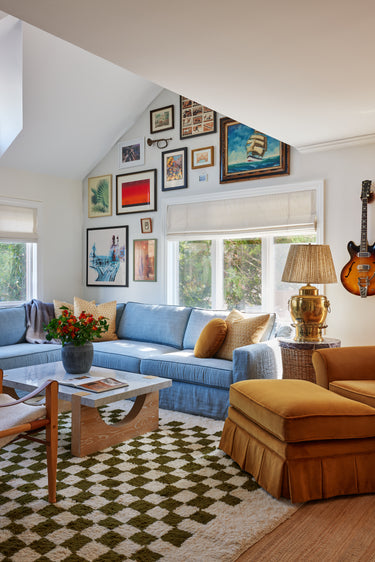 A bright, modern living room with a light blue sectional sofa, a mustard yellow ottoman, a black and white checkered rug, and a gallery wall of framed art on a white slanted ceiling. Custom Guitar hanger