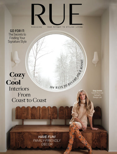 RUE magazine cover featuring a woman seated on a wooden bench in a room with a large round window showing a snowy outdoor scene. The cover includes prominent text such as 'RUE', 'Cozy + Cool Interiors', and 'Have Fun Family-Friendly Decor'.