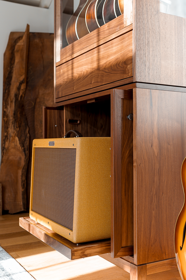 A vintage-style yellow guitar amplifier with a brown grille, tucked inside a wooden cabinet shelf in a room with warm sunlight and wood flooring.