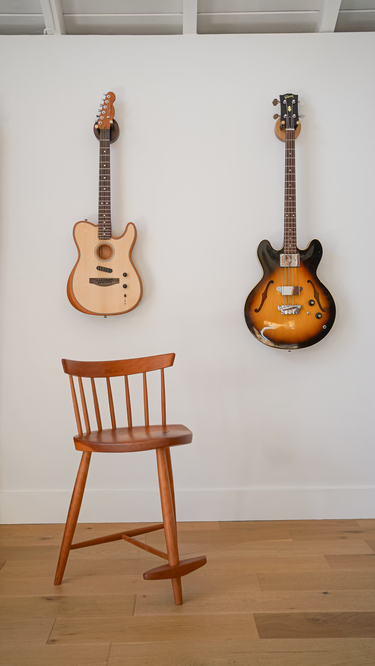 Two guitars hanging on a white wall with a wooden chair in front.